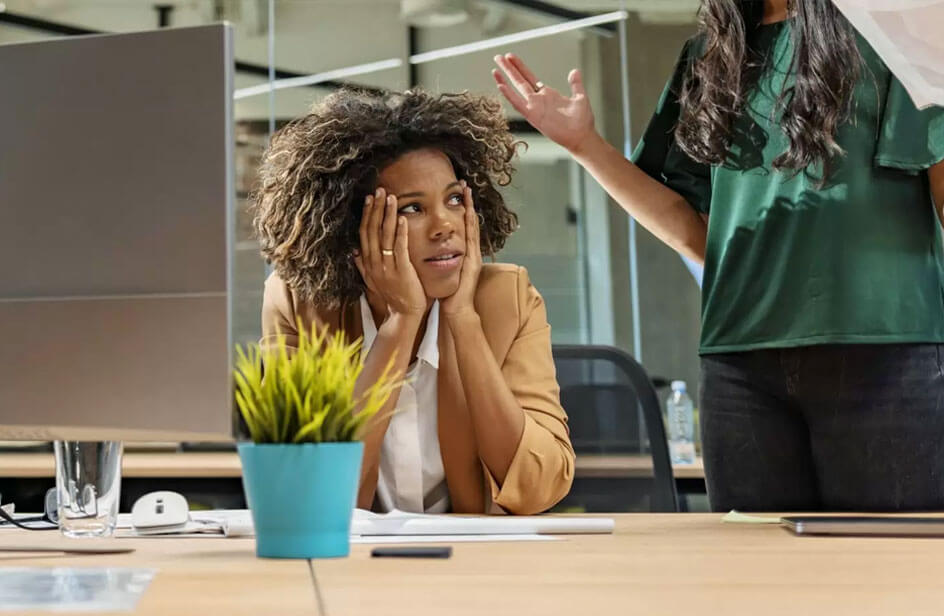 Women with hands on her face looks stressed as a colleague speaks to her partially out of frame - 4 Causes of Workplace Conflict